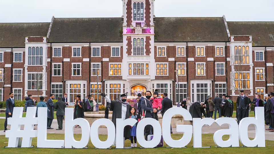 A photo of two adults and two children, standing behind large white letters that say #LboroGrad. The backdrop is the Hazlerigg Building.